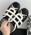 A pair of black and white platform caged sandals with white ribbon bows, held by a hand against a neutral gray background.
