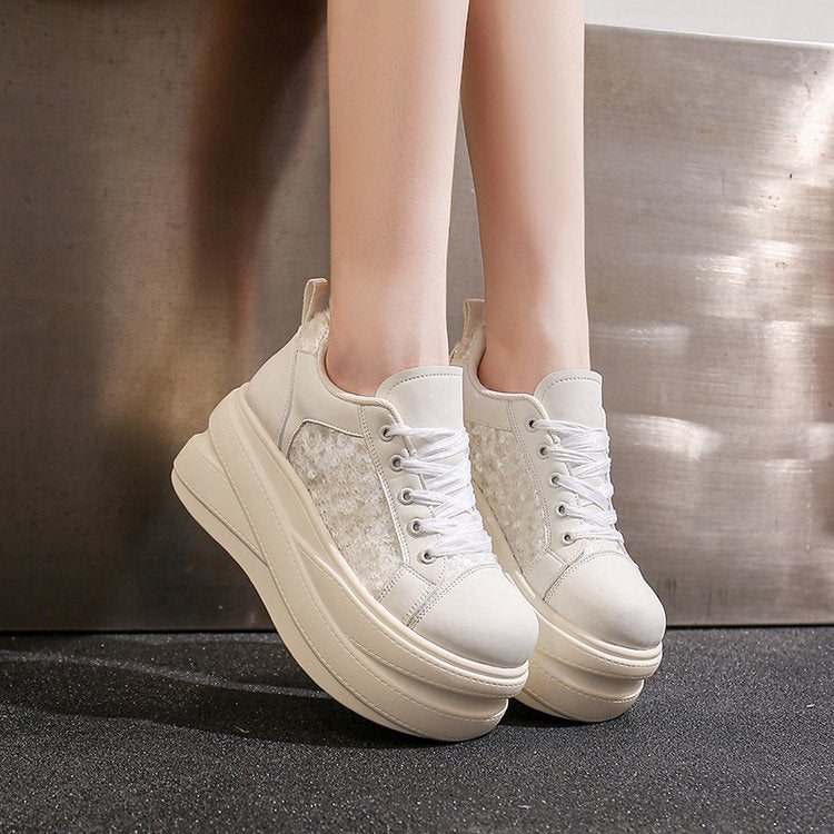 A pair of chunky platform sneakers in off-white and beige, worn by a model standing against a metallic wall.
