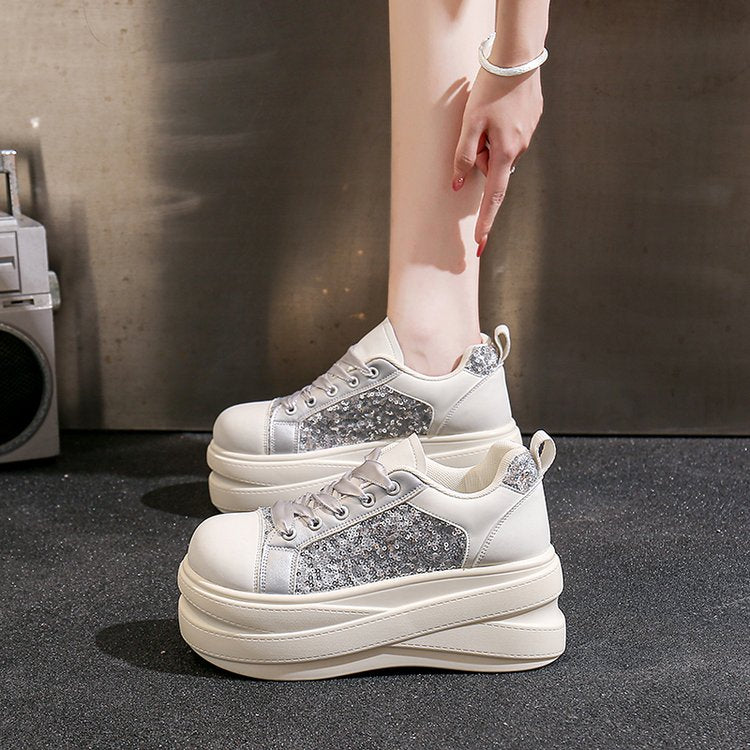 A pair of chunky platform sneakers in off-white and silver glitter, shown on a model's feet against a dark textured studio background.