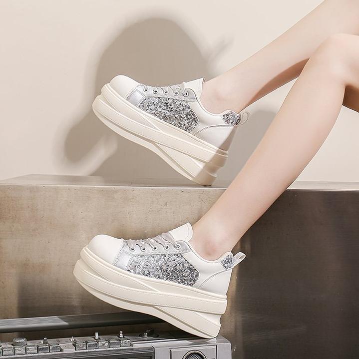 A pair of chunky platform sneakers with white leather and silver glitter panels, shown on a model's feet against a neutral studio background.