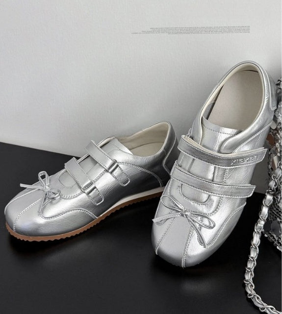A pair of metallic silver slip-on shoes with double hook-and-loop straps, decorative bows, and a tan outsole, displayed on a black surface against a light gray wall.