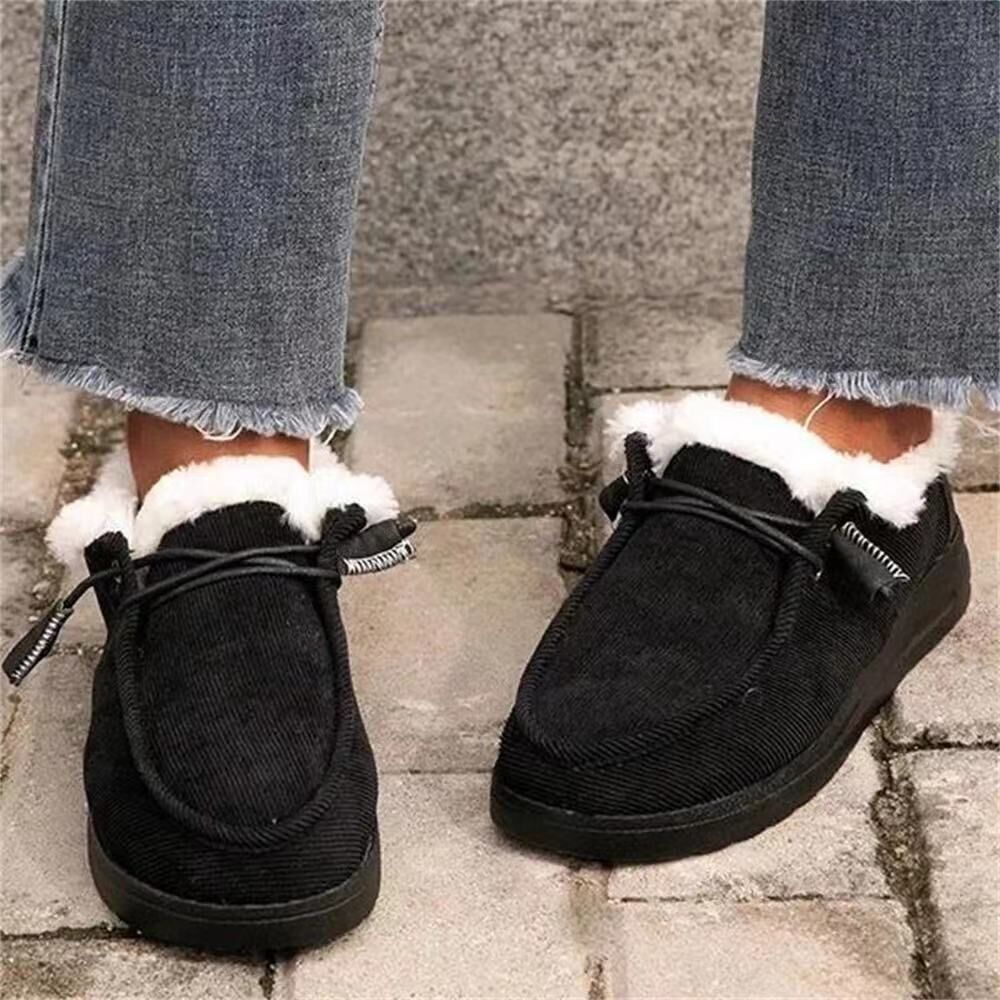 Black moccasin-style shoes with white fur trim worn with blue jeans on a stone pavement.